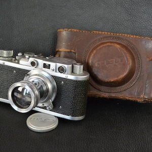 Puede incluir: Una cámara vintage plateada y negra con una lente grande, una tapa de lente y una funda de cuero marrón. La cámara tiene un cuerpo negro texturizado y detalles plateados. La funda de cuero está desgastada y muestra signos de edad.