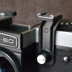 Puede incluir: Una cámara de película Kiev-60 negra y plateada con un accesorio de flash. La cámara tiene una cubierta de cuero negro y un cuerpo de metal plateado. El accesorio de flash es de plástico negro y tiene una perilla de metal plateado.