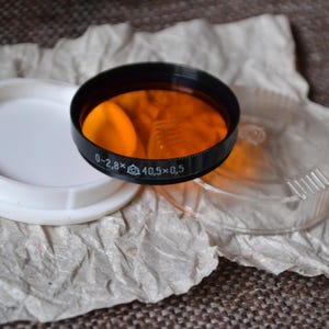Op de afbeelding: Een close-up van een zwart en oranje camera lensfilter. Het filter heeft een zwarte rand en een helder oranje midden. Het filter is gelabeld "0-2.8x 40,5 x 0,5".