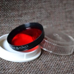 May include: A red filter for a camera lens, with a black rim and a white plastic case. The filter is labeled "40.5 x 0.5".