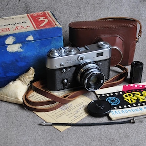 May include: A vintage FED 3 rangefinder camera with a silver and black body, accompanied by a brown leather case, a blue cardboard box with Cyrillic text, a black film canister, a lens cap, a shutter release cable, and a printed instruction manual.