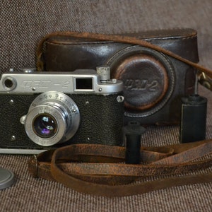 Puede incluir: Una cámara de película de 35 mm de color plateado y negro con una funda y una correa de cuero. La cámara tiene un objetivo con una tapa de color negro y plateado. La cámara está sobre una superficie marrón texturizada.