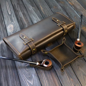 May include: A brown leather pipe case with two pipes and a small leather tray. The case has a flap with a buckle closure and is decorated with stitching and brass accents.