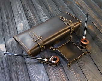 May include: A brown leather pipe case with two pipes and a small leather tray. The case has a flap with a buckle closure and is decorated with stitching and brass accents.