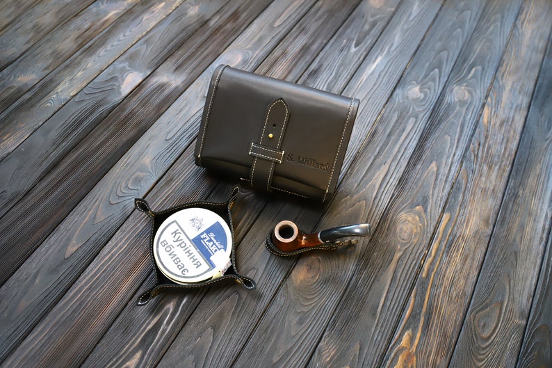Pu&ograve; includere: Una sacca per tabacco in pelle nera con una cinghia e il testo "S & Williams" in rilievo sul davanti. Una pipa in legno marrone e una scatola rotonda di tabacco sono anche raffigurate su una superficie in legno scuro.