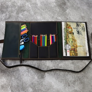 May include: Open black leather art portfolio with a green interior. The portfolio has multiple pockets for pencils, pens, and other art supplies. A sketchbook with a watercolor painting of a cityscape is inside the portfolio.