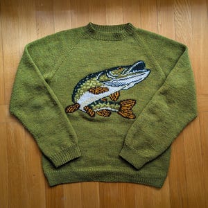 May include: A long-sleeved olive green knitted jumper featuring a large intarsia graphic of a fish on the chest. The fish design incorporates shades of white, dark green, and orange. The jumper has a ribbed crew neckline, cuffs, and hem.