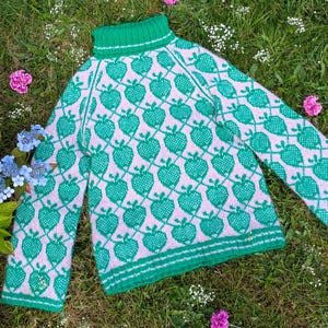 May include: A green and pink knitted sweater with a turtleneck collar. The sweater features a repeating pattern of green hearts on a pink background. The sleeves are long, and the sweater is lying on a bed of green grass.