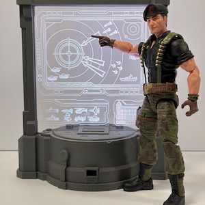 May include: An action figure of a military officer in a black uniform, camouflage pants, and black boots, pointing at a gray display screen with a target and military vehicle graphics. The figure wears a black beret and has a belt with ammunition.