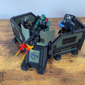 Könnte beinhalten: Eine grüne und schwarze Spielzeug-Militärbasis mit drei Spielzeugsoldaten. Die Basis hat ein Maschinengewehr mit orangefarbenen und roten Flammen. Die Soldaten tragen grüne und schwarze Uniformen und Helme.