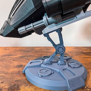 May include: A gray 3D printed model of a spaceship with a detailed design. The spaceship is mounted on a gray base with a circular platform and articulated legs.