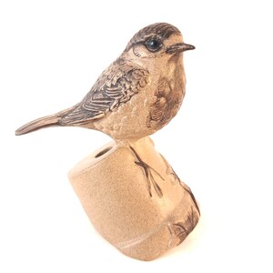 May include: A bronze-toned bird figurine perched on a textured, light-coloured stone-like base. The bird features detailed feather carvings, a dark eye, and a pointed beak. The base has a cylindrical shape with a small circular opening at the top.