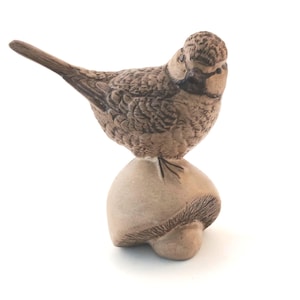 May include: A small ceramic figurine of a brown bird with detailed feather textures perched atop a smooth, light tan mushroom. The bird has a dark beak and eye markings, while the mushroom base features visible gill lines underneath its cap.