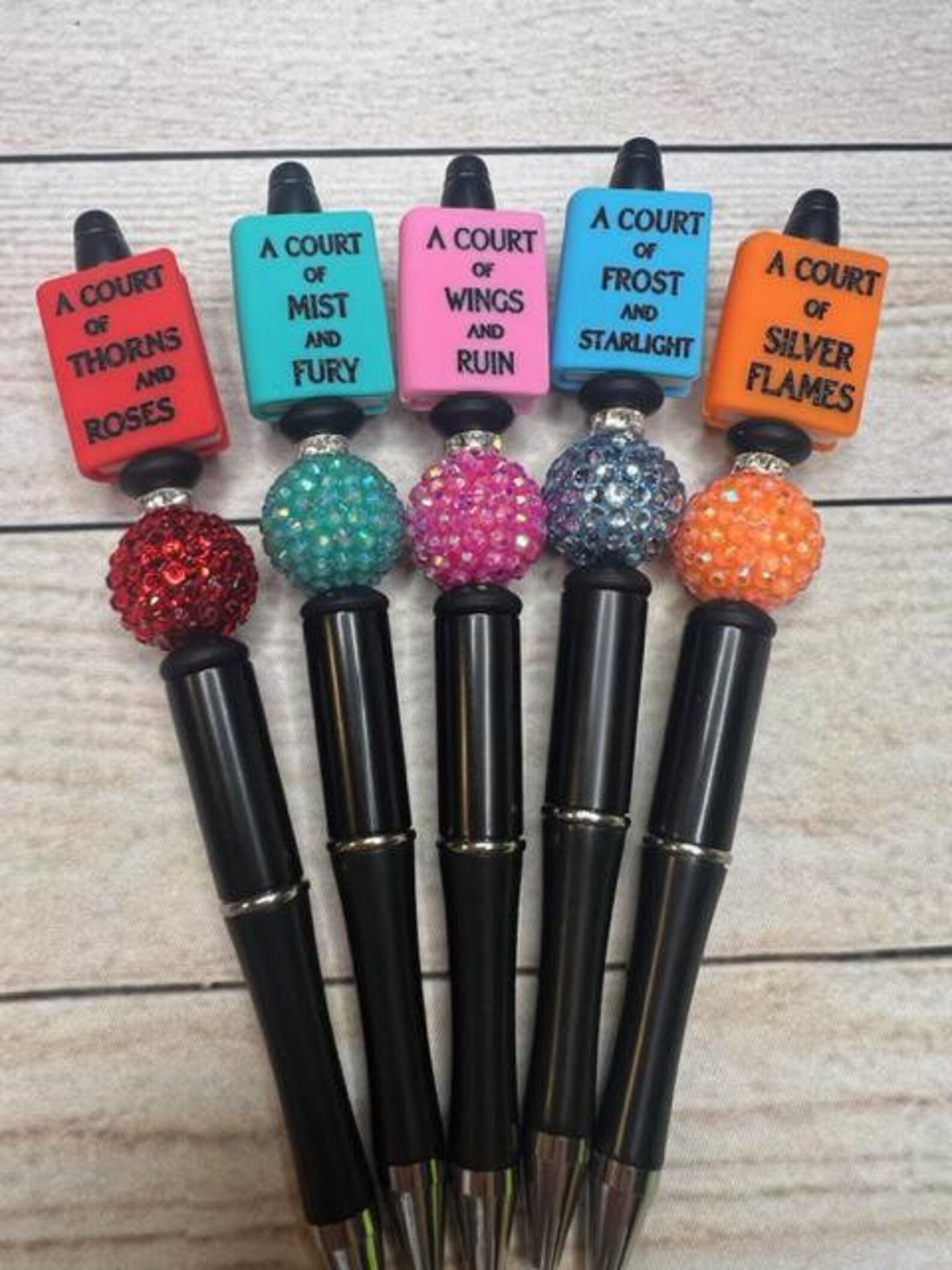 Five pens with decorations on the top that look like the five "A Court of Thorns and Roses" books in red, teal, pink, blue, and orange.