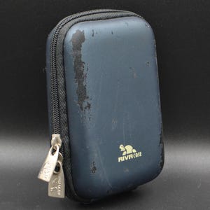 Puede incluir: Un estuche rectangular azul marino con cremallera negra y tiradores plateados. El estuche tiene un logotipo en la parte delantera y muestra signos de desgaste. Probablemente se utiliza para guardar aparatos electrónicos o accesorios.