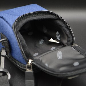 May include: A navy blue rectangular camera bag with a black interior and a zip closure. The bag has a black strap and a metal clasp. The interior has a white tag with the word "BIORA" printed on it.