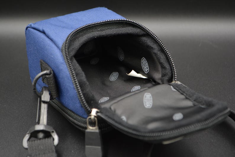 May include: A navy blue rectangular camera bag with a black interior and a zip closure. The bag has a black strap and a metal clasp. The interior has a white tag with the word "BIORA" printed on it.