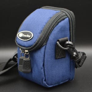 May include: A navy blue EverStar camera bag with a black zip and strap. The bag features a small oval logo and a black clasp. The bag is designed to hold a camera and accessories, and is approximately 15 cm high.