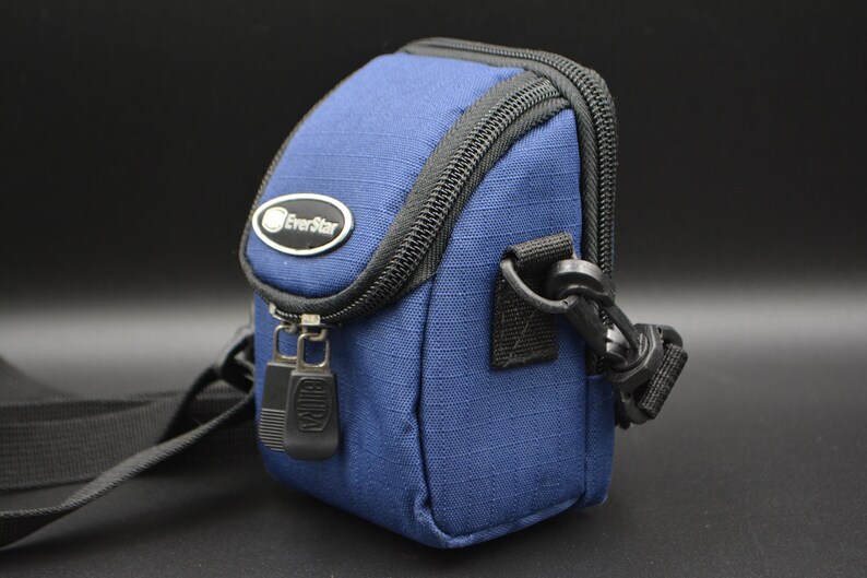 May include: A navy blue EverStar camera bag with a black zip and strap. The bag features a small oval logo and a black clasp. The bag is designed to hold a camera and accessories, and is approximately 15 cm high.