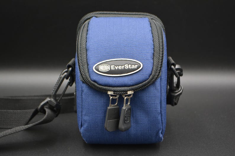 May include: A blue EverStar camera bag with a black zip and strap. The bag is rectangular with rounded edges and a black zip. The EverStar logo is on the front of the bag.