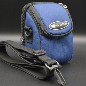 May include: A blue camera bag with a black strap. The bag has a zippered closure and a small metal emblem that reads "EverStar". The strap is adjustable and has a metal clasp.
