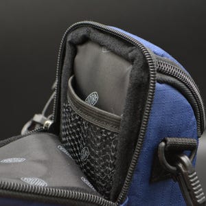 May include: A navy blue camera bag with a black interior and a mesh pocket. The bag has a black zip and a black strap attachment. The interior has a logo. The bag is open, revealing the inside.