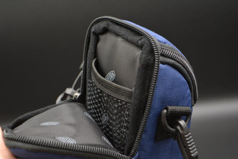 May include: A navy blue camera bag with a black interior and a mesh pocket. The bag has a black zip and a black strap attachment. The interior has a logo. The bag is open, revealing the inside.