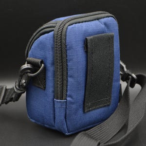 May include: A small, navy blue camera bag with a black zip, strap, and clip. The bag has a rectangular shape and a black strap on the back. The bag is designed to be worn over the shoulder.