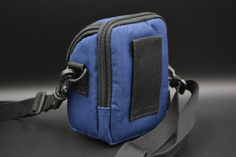 May include: A small, navy blue camera bag with a black zip, strap, and clip. The bag has a rectangular shape and a black strap on the back. The bag is designed to be worn over the shoulder.