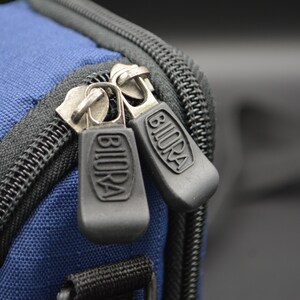 May include: Close-up of a blue fabric bag with black zippers. The zipper pulls are dark grey with the word "BIURA" embossed on them. The bag has a black zipper and trim.