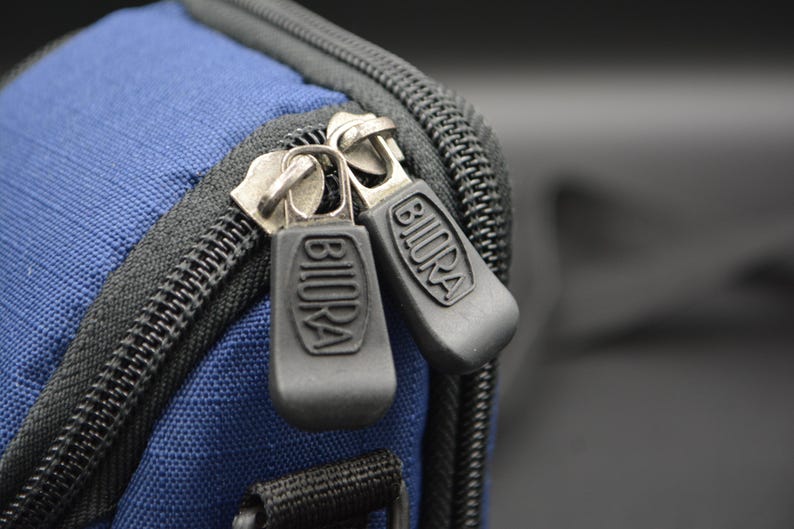 May include: Close-up of a blue fabric bag with black zippers. The zipper pulls are dark grey with the word "BIURA" embossed on them. The bag has a black zipper and trim.