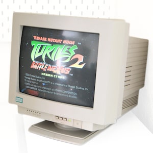 Peut inclure: Un moniteur d'ordinateur beige vintage affichant le jeu Teenage Mutant Ninja Turtles 2: Battle Nexus. L'écran montre le titre du jeu en vert, jaune et rouge, avec un fond étoilé. Le moniteur porte le logo Siemens.