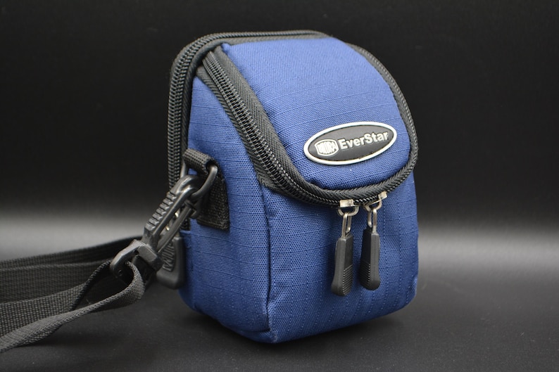 May include: A navy blue camera bag with a black zip and strap. The bag has a curved top and an EverStar logo. Designed to hold a camera and accessories. The bag is approximately 15 cm high.