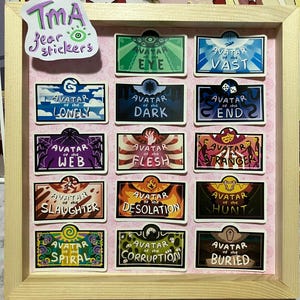 May include: A wooden framed display of "TMA fear stickers". The stickers are rectangular with various designs and text, including "Avatar of the Eye", "Avatar of the Vast", and "Avatar of the Lonely". The background is pink.