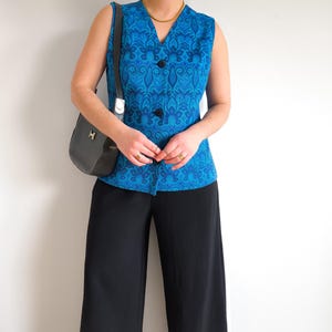 Könnte beinhalten: Eine ärmellose blaue Weste mit floralem Muster und schwarzen Knöpfen. Die Weste wird mit einer schwarzen Hose mit weitem Bein und einer schwarzen Umhängetasche kombiniert. Eine goldene Halskette und Ringe vervollständigen das Outfit.