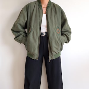 Könnte beinhalten: Olivgrüne Bomberjacke mit geripptem Kragen, Bündchen und Saum. Die Jacke hat einen Frontreißverschluss und zwei Vordertaschen. Rautenartige Aufnäher mit einem orangefarbenen und braunen geometrischen Design befinden sich auf der Brust. Schwarze Hose.