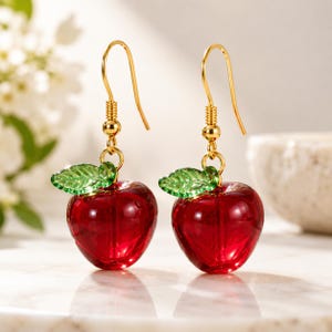 May include: A pair of dangle earrings featuring translucent red glass apple charms with small green glass leaves. The charms are suspended from gold-toned metal fishhook wires, creating a bright and reflective appearance for these fruit-themed accessories.