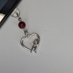 May include: A silver-toned charm featuring a heart-shaped pendant with a small cherub figure inside. A dark red bead is connected to the charm by a silver chain. The charm is attached to a phone.