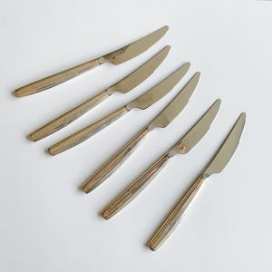 Op de afbeelding: Zes vintage roestvrijstalen messen met een zilveren lemmet en een goudkleurig handvat. De messen zijn diagonaal op een wit oppervlak geplaatst. De messen zijn waarschijnlijk voor een formele eetsituatie.