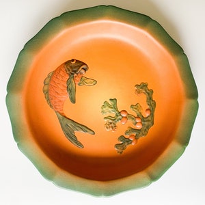 May include: A round orange ceramic dish with a green rim. The dish features a raised design of a fish and a leafy branch with berries.