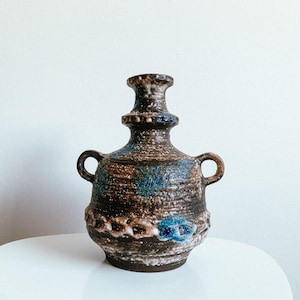 Op de afbeelding: Een vintage keramische vaas met een getextureerd oppervlak. De vaas heeft een bruin en blauw kleurenschema met twee kleine handvatten. Het ontwerp omvat decoratieve elementen en een unieke vorm, geschikt voor interieurdecoratie.