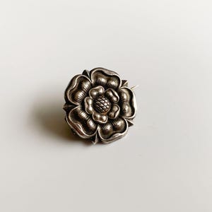 Puede incluir: Un broche de plata con forma de rosa y superficie texturizada.