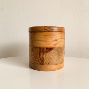 Può includere: Un contenitore cilindrico costruito con più pezzi di legno marrone chiaro con venature visibili. L'esterno presenta un design a mosaico di segmenti di legno orizzontali e verticali uniti insieme per formare un solido recipiente dalle pareti lisce.