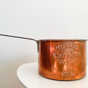 Può includere: Una casseruola vintage in rame con un lungo manico. La pentola presenta testo in rilievo e l'anno 1931 sul lato. Il rame ha un colore marrone-rossastro caldo e mostra segni di età.