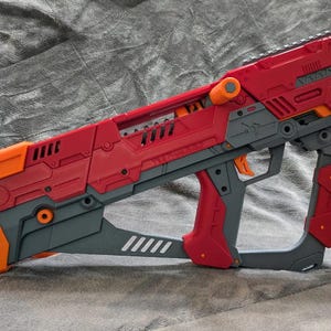 以下が含まれることがあります： グレーのハンドルが付いた赤とオレンジの玩具の銃。銃は多くのディテールを持つ未来的なデザインです。