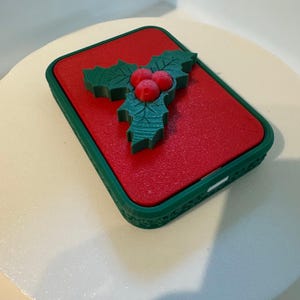 May include: A red rectangular case with a green border and a holly design on top. The holly features green leaves and three red berries, creating a festive, seasonal theme. The case has a textured surface.