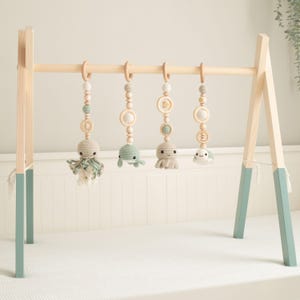 May include: A wooden baby play gym with four hanging crochet sea creatures: an octopus, a whale, a sea turtle, and a small fish. The wooden frame has light blue accents on the legs and natural wood tones throughout. The toys are suspended from wooden rings.