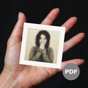 May include: A digital cross stitch pattern of a person with long black hair wearing a green and black jacket. The pattern is in a white frame and is being held in a person's hand. The word "PDF" is written in grey on a grey circle.