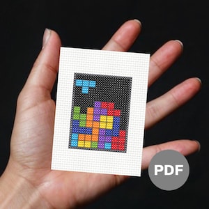 Image of a mini cross stitch of an 80s puzzle video game. It is held in an open palm and has raw Aida edges.