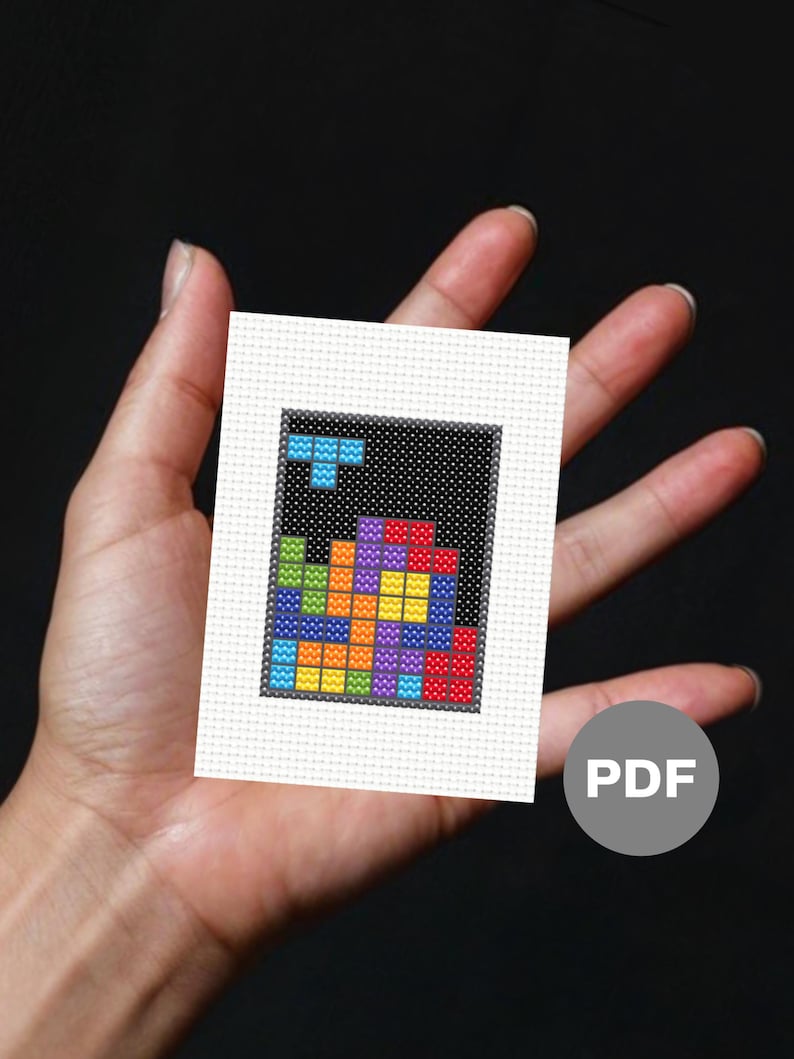 Image of a mini cross stitch of an 80s puzzle video game. It is held in an open palm and has raw Aida edges.